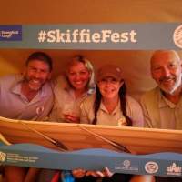 Four visitors pose to take a photograph, they hold up a Skiffie Fest frame which has a skiff on the bottom