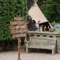 A wooden signpost stands beside a bench, a tipi peeks behind a hedge in the background