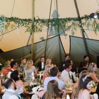 Guest sit and listen to a speech, foliage and flowers decorate the beams inside the wedding Tipi