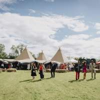Guests stand outside talking with a four tipi Wedding reception on an open field, square hay-bales sit in front