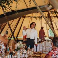 The groom smiles and holds his drink up in front of guests inside of the tipi wedding tent