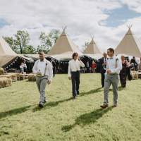 The groom and groomsmen walk with the tipi wedding reception standing in the background