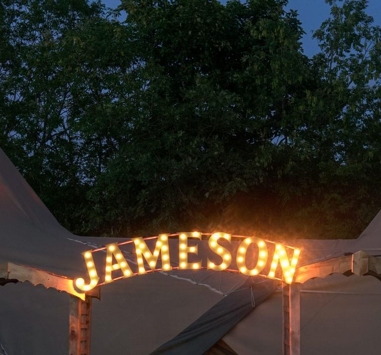 Two festival tipis stand with a double archway in front with Jameson light sign decorating the top