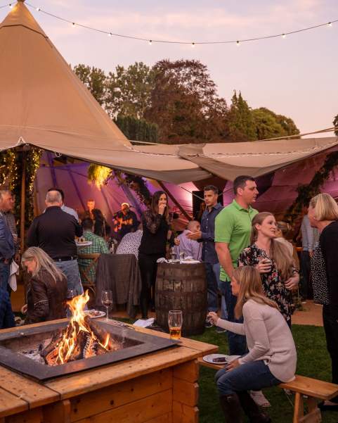 Corporate guests sit at a firepit and chat, open sided Tipis are behind them