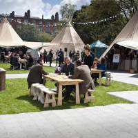 Tipis are joined in a ring, with bunting decoration, people sit at picnic benches in the foreground