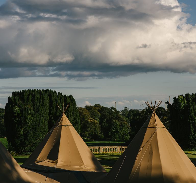 Three Tipis stand in the forefront with the grounds of Narrow Water Castle in the background and a cloudy bright sky