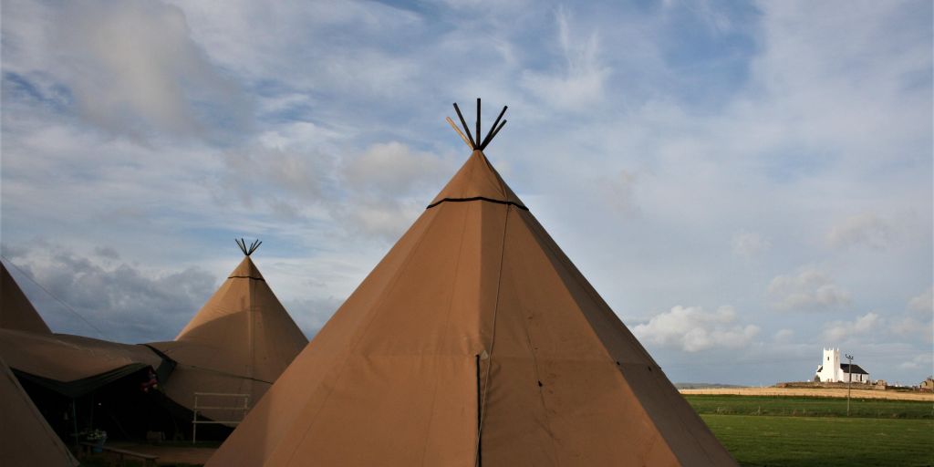Tipi tops stand in front of a cloudy blue sky with a white church and green fields.