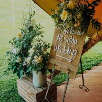 A wooden sign saying welcome to our wedding sits on an easel, flower bouquets sit in metal buckets beside it