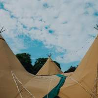 Three Tipis are joined together, overlapping canvases and festoon lighting, a bright blue cloudy sky is in the background