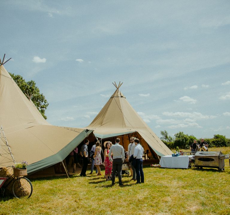 Guest arrive at a three tipi wedding set up, tables and bbq's stand outside beside the tipi entrance