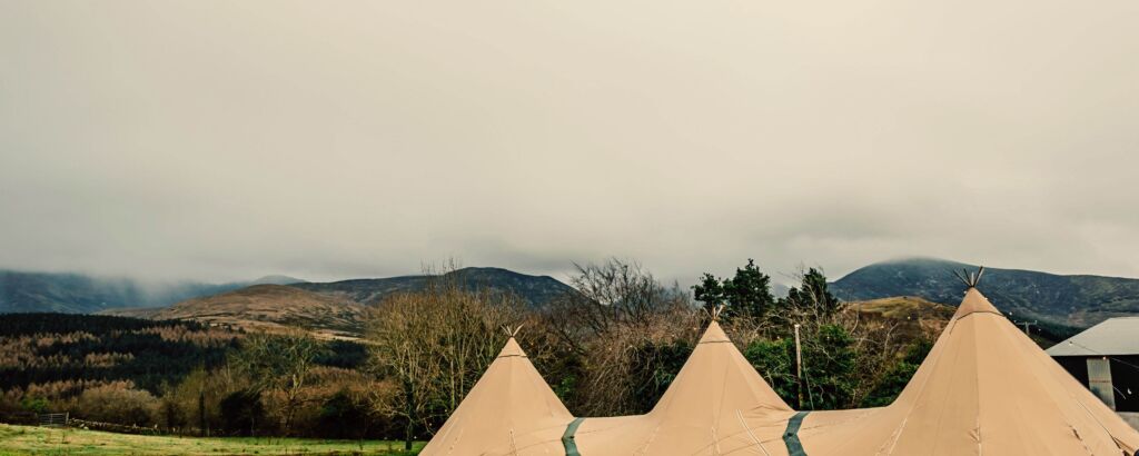 Three Tipis are setup in a field with beautiful irish countryside in the background