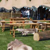 A six tipi festival fair set up with picnic benches and reindeer skins on logs sitting in front on the green
