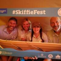 Four visitors pose to take a photograph, they hold up a Skiffie Fest frame which has a skiff on the bottom
