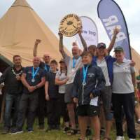 Rowers cheer and pose for a photo, holding up a circular award, Two tipis are behind them