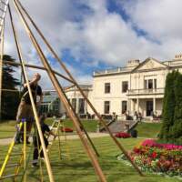 Two magnakata crew set up the poles of a tipi, a white mansion with lovely green gardens are in the background