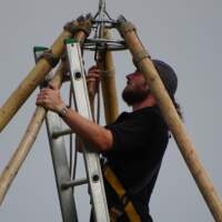 A magnakata crew member is on a ladder fitting the central tipi wooden poles together