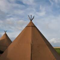 Tipi tops stand in front of a cloudy blue sky with a white church and green fields.
