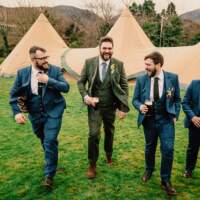 The groom and groomsmen smile and laugh, holding their drinks as they walk, three tipis are behind them