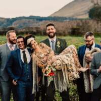 The groom carries the bride in his arms, surrounded by the groomsmen all suited up