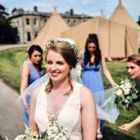 The bride walks with her bridesmaids lifting her veil, Tipis and the family estate are in the background