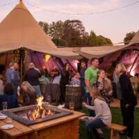 Corporate guests sit at a firepit and chat, open sided Tipis are behind them