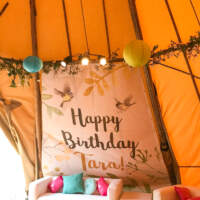 A personalised birthday banner decorates a panel of a tipi, a white sofa set and colourful paper lanterns decorate the tipi