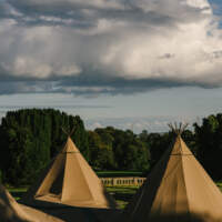 Three Tipis stand in the forefront with the grounds of Narrow Water Castle in the background and a cloudy bright sky
