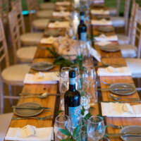 A long wooden table is decorated with place setting, simple foliage and a bottle of wine