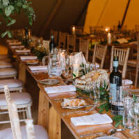 A row of long wooden tables is decorated with foliage, candles, a basket of bread and a bottle of wine sits in the foreground