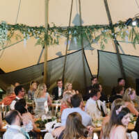 Guest sit and listen to a speech, foliage and flowers decorate the beams inside the wedding Tipi