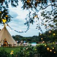 Fairylights decorating the trees frame a lovely wedding tipi setup with chairs and benches outside in a walled garden