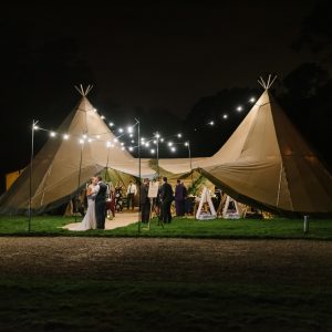 Bride & groom kiss under festoon lighting at night in the entrance of their tipi wedding reception