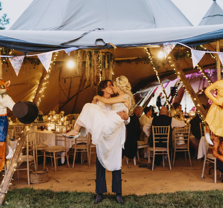 Groom holds the bride as they kiss at the entrance of a tipi decorated by bunting and Fox paper maché of themselves