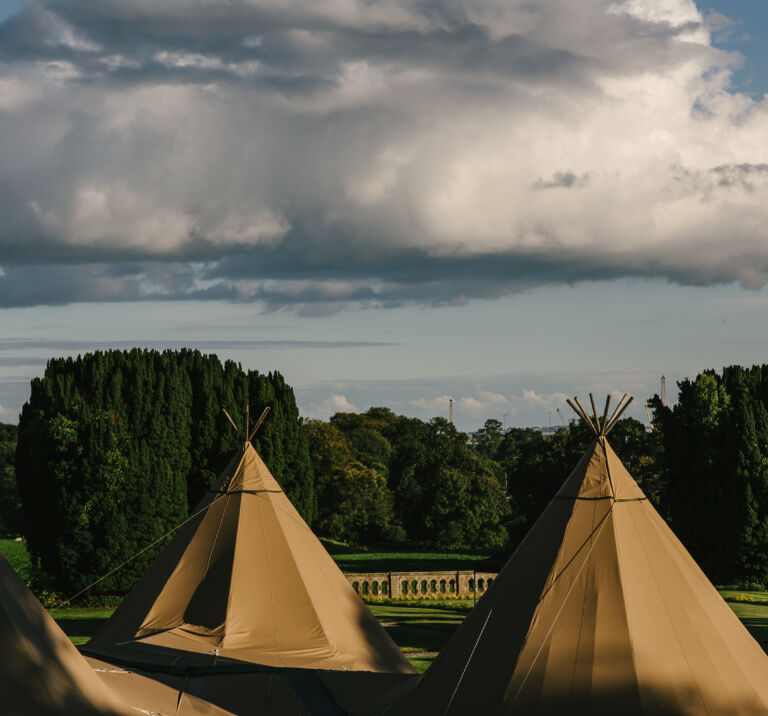 Three Tipis stand in the forefront with the grounds of Narrow Water Castle in the background and a cloudy bright sky