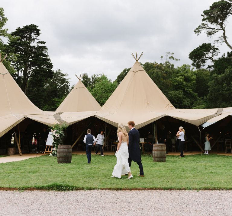 Bride & groom walk together in front of four linked tipis set up on the grounds of Mount Stewart