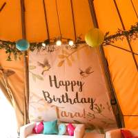 A personalised birthday banner decorates a panel of a tipi, a white sofa set and colourful paper lanterns decorate the tipi