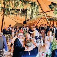 Wedding guests celebrate at their tables, the wedding Tipis are decorated in lush hanging foliage and flowers