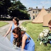Three bridesmaids dressed in blue carry the brides veil, Tipis, a catering tent and the family estate are in the background