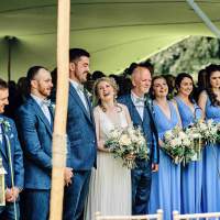 Bride and groom and their wedding party stand in a row during their wedding ceremony underneath a stretch tent