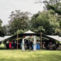 A wedding ceremony takes place under a stretch tent, a wooden arch decorated in white fabric stand in the forefront