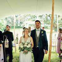 The bride and groom walk through their guests after their outdoor ceremony under a stretch tent, lanterns line the aisle