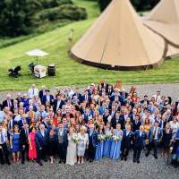 Wedding guests gather to pose for a photograph, an ice cream trolley and wedding tipis are behind them