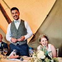 The groom stands to speak, the bride sits beside him laughing inside their wedding Tipis