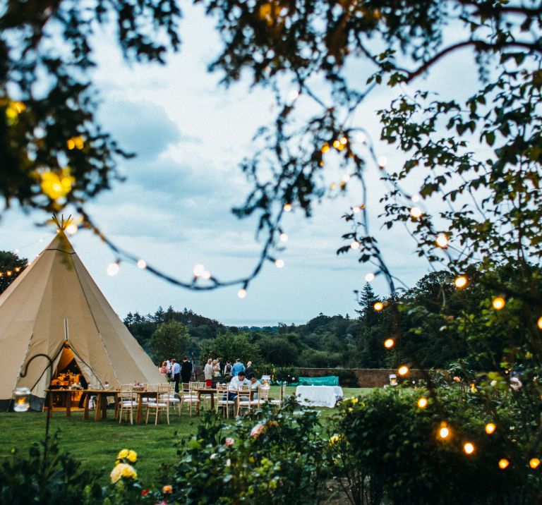 Fairylights decorating the trees frame a lovely tipi party setup with chairs and benches outside in a walled garden