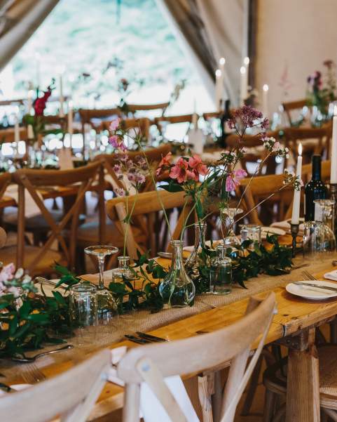 Rows of long log tables are decorated with wildflowers and foliage with gold candlesticks and tapered dinner candles inside a tipi