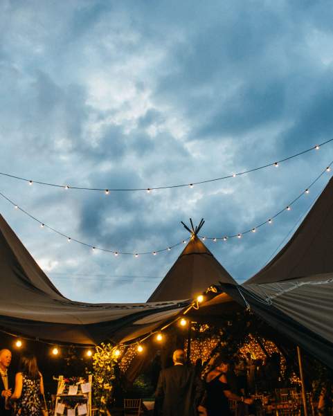 Three tipis are joined their central poles highlighted by the sky, festoon lighting hangs creating a cosy atmosphere