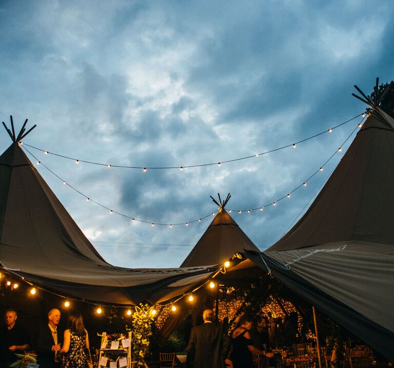 Three tipis are joined their central poles highlighted by the sky, festoon lighting hangs creating a cosy atmosphere