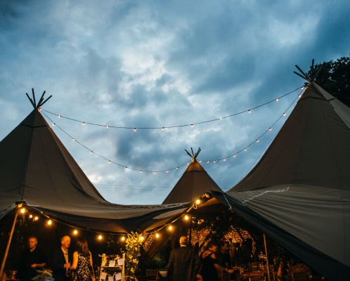 Three tipis are joined their central poles highlighted by the sky, festoon lighting hangs creating a cosy atmosphere