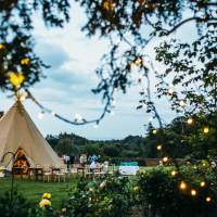 Fairylights decorating the trees frame a lovely wedding tipi setup with chairs and benches outside in a walled garden