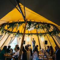 Guests sit inside a tipi decorated with fairylit panels and natural foliage crossbeams, the best man gives a speech